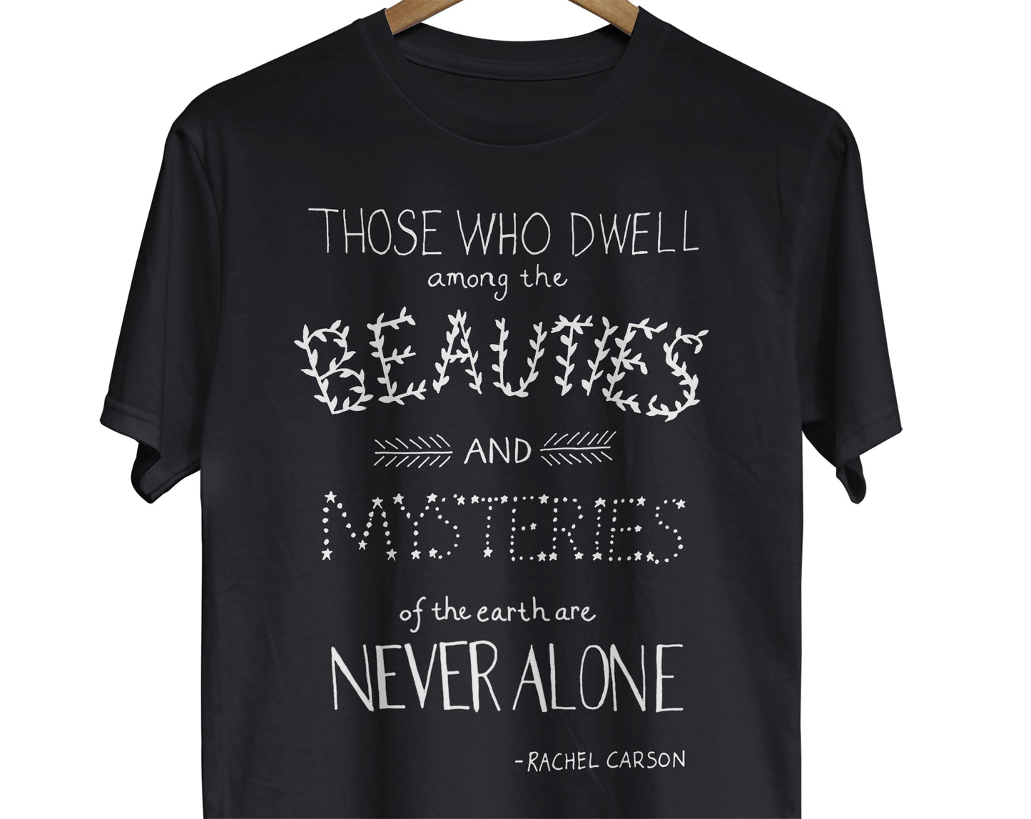 A t-shirt featuring a hand-lettered quote by Rachel Carson. The quote reads, "Those who dwell among the beauties and mysteries of the earth are never alone."